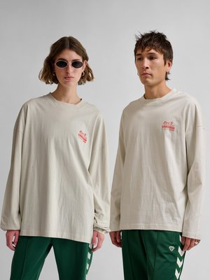 Dos jóvenes adultos con camisetas beige de manga larga con texto rojo y pantalones verdes con rayas blancas en forma de chevrón están de pie frente a un fondo liso.