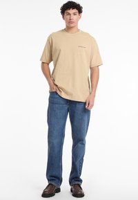 Bēšas kokvilnas t-krekls ar apaļu kakla un īsām piedurknēm, ar mazu melnu "Guess Jeans" logo, kopā ar ziliem brīvi pieguļošiem džinsiem.