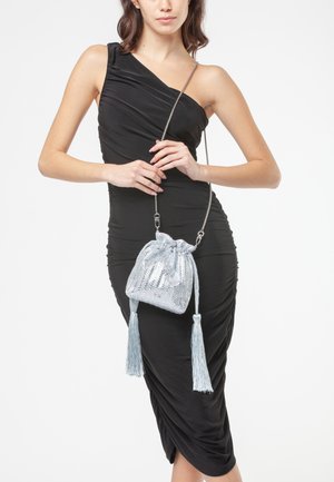 Bolso de cuerda plateado con acabado texturizado, adornado con dos borlas largas y una correa de cadena metálica, sostenido contra un vestido negro.