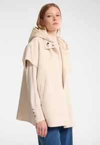 Giovane donna che indossa una cappotto senza maniche color crema con cappuccio sopra un maglione a maniche lunghe e jeans blu, in piedi davanti a uno sfondo bianco semplice.