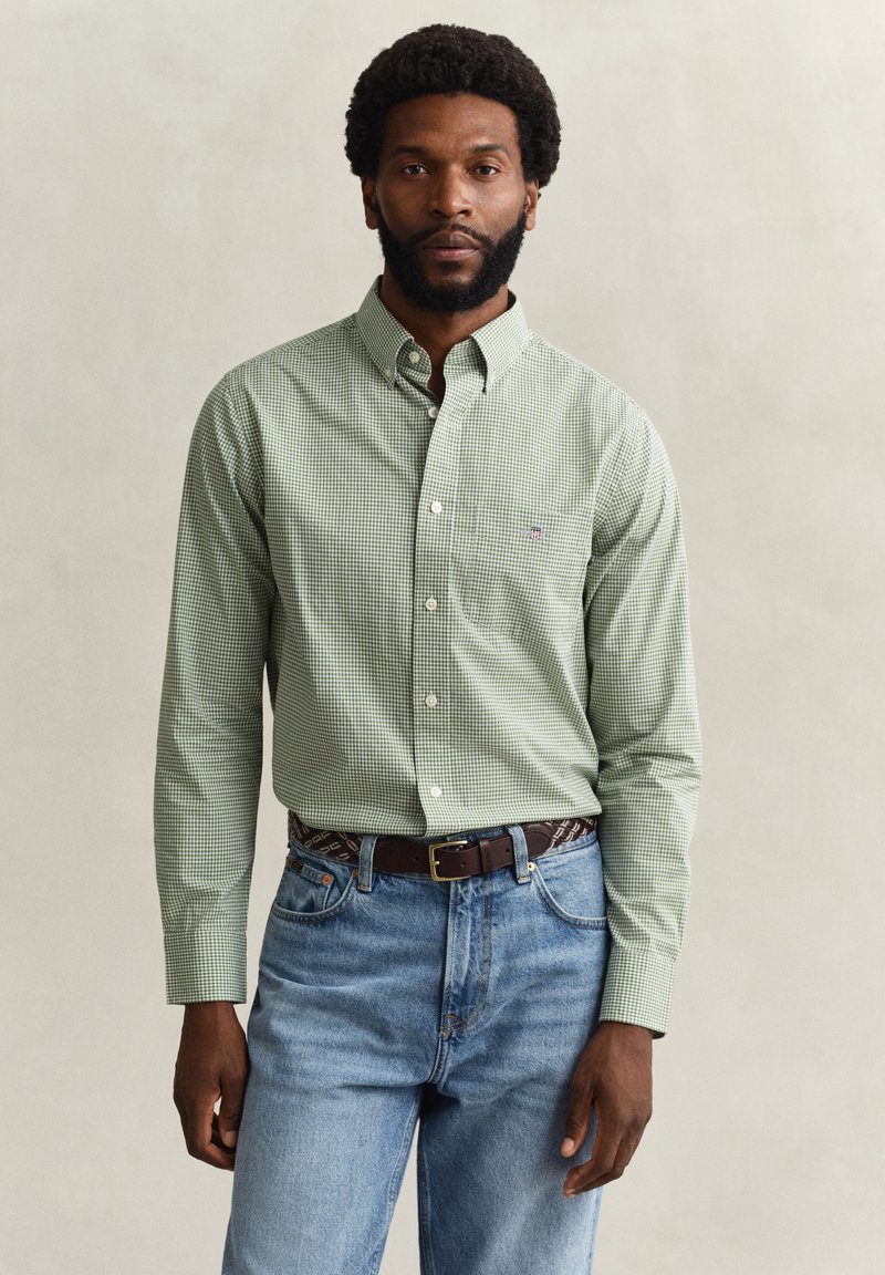 Homme portant une chemise à carreaux verte boutonnée, rentrée dans un jean bleu avec une ceinture marron, debout devant un fond clair uni.