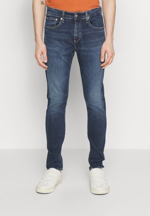 Jeans skinny blu scuro in denim, con design a cinque tasche, accenti sbiaditi e cuciture a contrasto, abbinati a sneaker bianche.