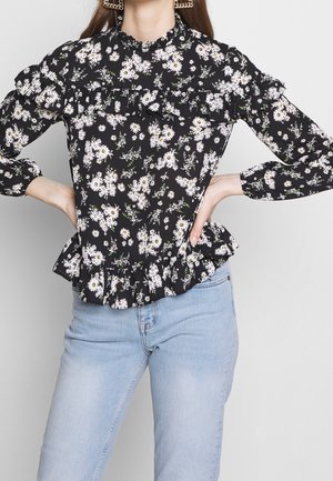 Zwart bloesje met witte madeliefjes, geplooide kraag en manchet, gelaagd zoom, gemaakt van lichtgewicht stof, gecombineerd met lichtblauwe jeans.