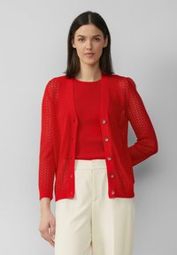 Roter gestrickter Cardigan mit Zickzackmuster, Knopfverschluss und langen Ärmeln, über einem roten gerippten Oberteil und cremfarbigen Hosen getragen.