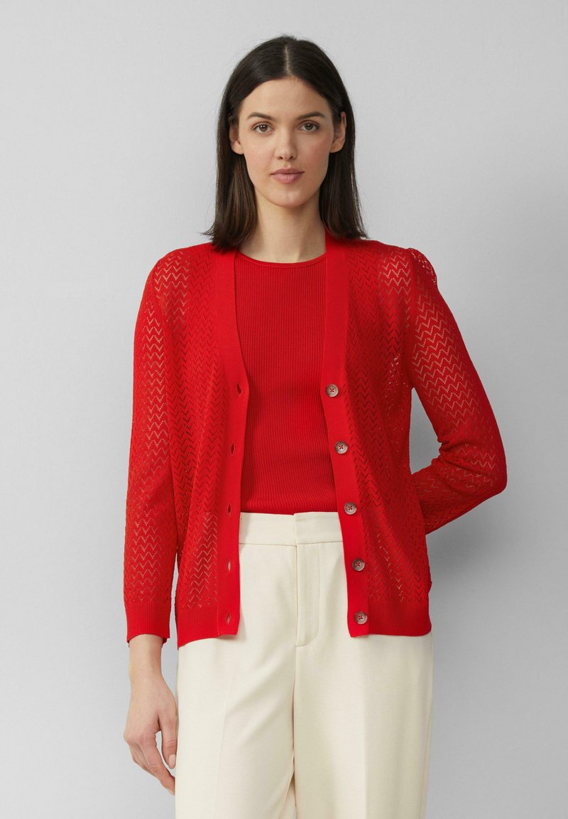 Roter gestrickter Cardigan mit Zickzackmuster, Knopfverschluss und langen Ärmeln, über einem roten gerippten Oberteil und cremfarbigen Hosen getragen.