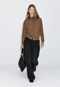 Cardigan lavorato a maglia marrone con colletto alto e chiusura anteriore con bottoni, indossato con pantaloni a gamba larga neri e tenendo una borsa in pelle nera.