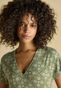 Mujer joven con cabello rizado, vestida con una blusa verde con estampado y cuello en V, y un collar con un colgante en forma de estrella dorada, sobre un fondo beige.