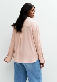Blusa rosa chiaro a maniche lunghe in tessuto morbido, caratterizzata da un colletto classico e polsini con bottoni, abbinata a jeans azzurri a vestibilità rilassata.