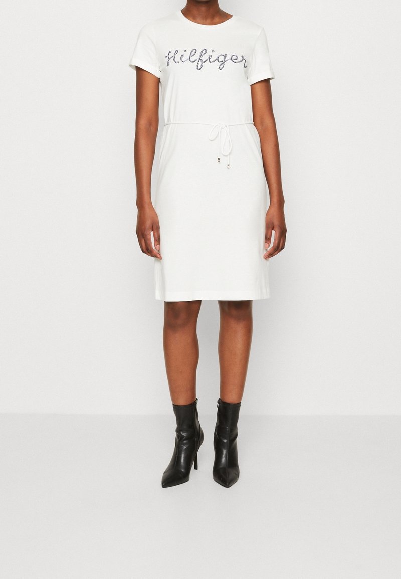 White t-shirt dress featuring a drawstring waist and "Hilfiger" logo in a contrasting color. Paired with black ankle boots.