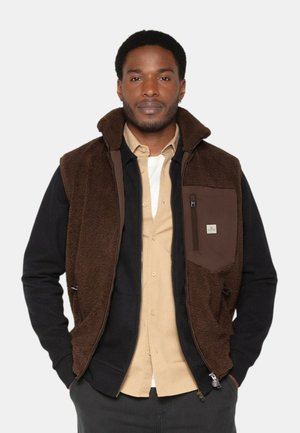 Gilet en polaire marron avec une couche de base noire, doté d'une poche poitrine zippée et d'une finition douce et texturée. Chemise à boutons en dessous.