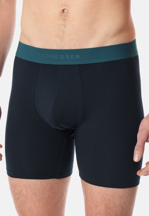 Schwarze Baumwollboxershorts mit einem breiten, dunkelblauen Bund, der den Markennamen "SCHIESSER" in erhabener Schrift trägt. Glatte Textur, enganliegender Schnitt.