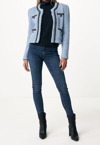 Veste texturée bleu clair avec des bordures noires et deux poches avant, portée sur un col roulé noir et associée à un jean skinny bleu et des bottines noires.