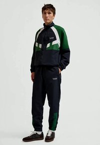 Jovem de pé, vestido com um fato de treino Hummel preto e verde e ténis pretos com atacadores brancos, contra um fundo claro e liso.