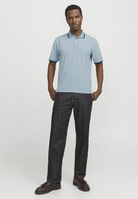 Ljusblå polo med marinblå detaljer, kombinerad med mörk denimjeans. Modellen står självsäkert i bruna skor. En enkel, klassisk design.