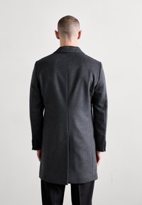 Cappotto di lana grigio con silhouette lunga, spacchetto singolo sul retro e dettagli con bottoni sulle maniche, abbinato a pantaloni neri sartoriali.