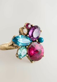 Aukso atspalvio žiedas, papuoštas multicolor akmenimis: gyvybingais rožiniais, violetiniais, turkio ir mėlynais akmenimis įvairių formų ir dydžių, su tekstūruota juosta.