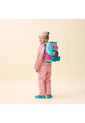 Blondes Kind in rosa Outfit schaut zurück, trägt einen türkis-rosa Eulen-Rucksack und passende türkisblaue Schuhe vor beigem Hintergrund.