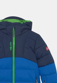 Veste matelassée avec un design en blocs de couleurs bleu et marine. Présente une fermeture éclair verte, une capuche et un petit patch du drapeau norvégien sur la manche.
