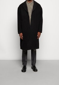 Ein Mann trägt einen langen schwarzen Mantel über einem beigen und schwarz karierten Hemd, ausgewaschenen schwarzen Jeans und schwarzen Schnürschuhen und steht vor einem weißen Hintergrund.