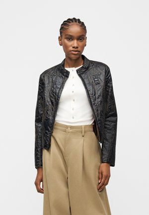 Moteris dėvi juodą siūtą striukę ant baltos su sagomis palaidinės ir aukšto liemens plačias smėlio spalvos kelnes, stovinti priešais baltą foną.