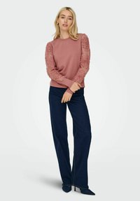 Felpa rosa con maniche a sbuffo floreali testurizzate, abbinata a jeans a gamba larga blu scuro e tacchi a punta neri, su uno sfondo chiaro.