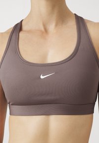 Soutien-gorge de sport athlétique marron en tissu lisse, avec un décolleté arrondi et un logo Nike blanc à l'avant.