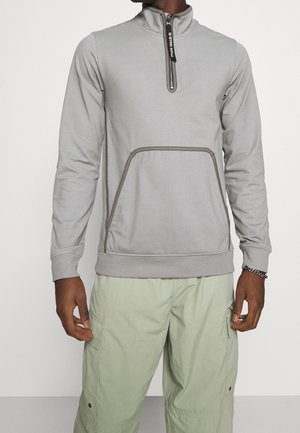 Homme portant un sweat-shirt gris clair à fermeture éclair au tiers avec poche avant et pantalon cargo vert clair sur fond blanc.