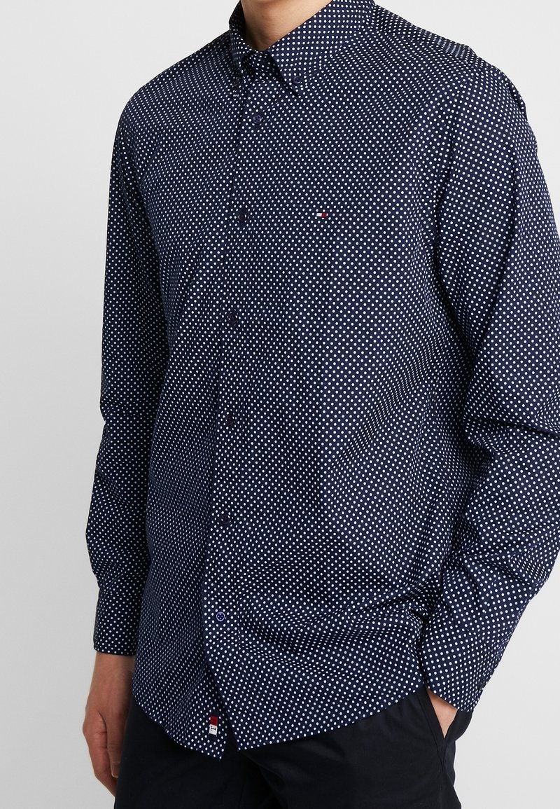 Camisa azul de botones con lunares blancos, cuello, mangas largas y un pequeño detalle de logo en el dobladillo. Hecha de una tela ligera.
