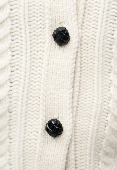 Cardigan tricoté blanc avec une texture côtelée épaisse et deux boutons noirs tissés pour la fermeture. Le design global est simple et harmonieux.
