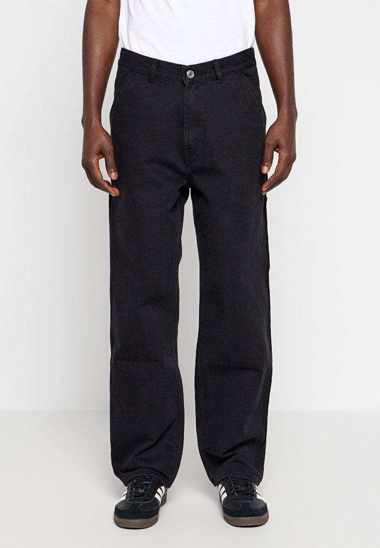 Pantalones de mezclilla negros con un corte recto, costuras visibles, cremallera y cierre de botón. Llevados con una camiseta blanca y zapatillas negras.