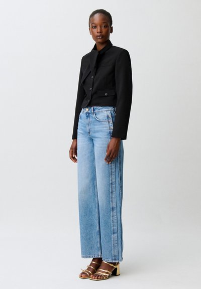 Donna che indossa un blazer nero corto, jeans azzurri a gamba larga con bottoni laterali e sandali dorati con tacco largo e cinturini, in piedi davanti a uno sfondo semplice.