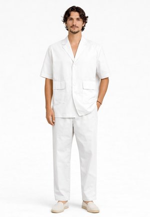 Uomo in piedi che indossa blazer bianco a maniche corte, pantaloni bianchi abbinati e scarpe espadrillas slip-on su sfondo bianco uniforme.