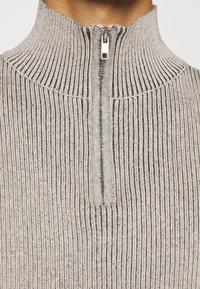 Maglione grigio a coste con collo alto e mezza cerniera. Il tessuto presenta un motivo a maglia strutturata con strisce scure, che ne evidenziano profondità e dimensione.