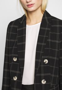 Svart rutigt kavaj med vita linjer, dubbelknäppt design, metallknappar och ett mjukt, texturerat tyg. Bärs över en vit topp.