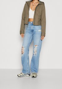 Kvinna i en cropped olivgrön jacka, vit knäppt crop top, ljusblå slitna vida jeans och beige sneakers som står framför en enkel bakgrund.