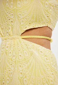 Rochie galbenă cu broderie florală, având laterale decupate, șnur împletit, țesătură texturată și modele circulare.