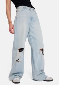 Jeans azzurri chiari a gamba larga con strappi sfrangiati alle ginocchia e vita alta. Presentano un classico design a cinque tasche e una leggera texture sfumata.