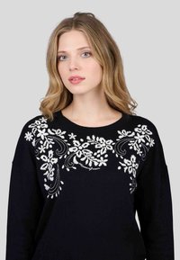 Pull noir avec broderie florale blanche sur la poitrine, doté d'un col rond et de manches amples. Texture lisse.