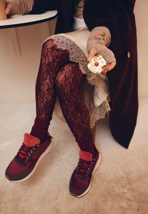 Scarpe da ginnastica color bordeaux con dettagli in pelliccia rossa, abbinate a leggings neri con pizzo. Una pasta decorativa è tenuta in una mano. Pavimento chiaro.