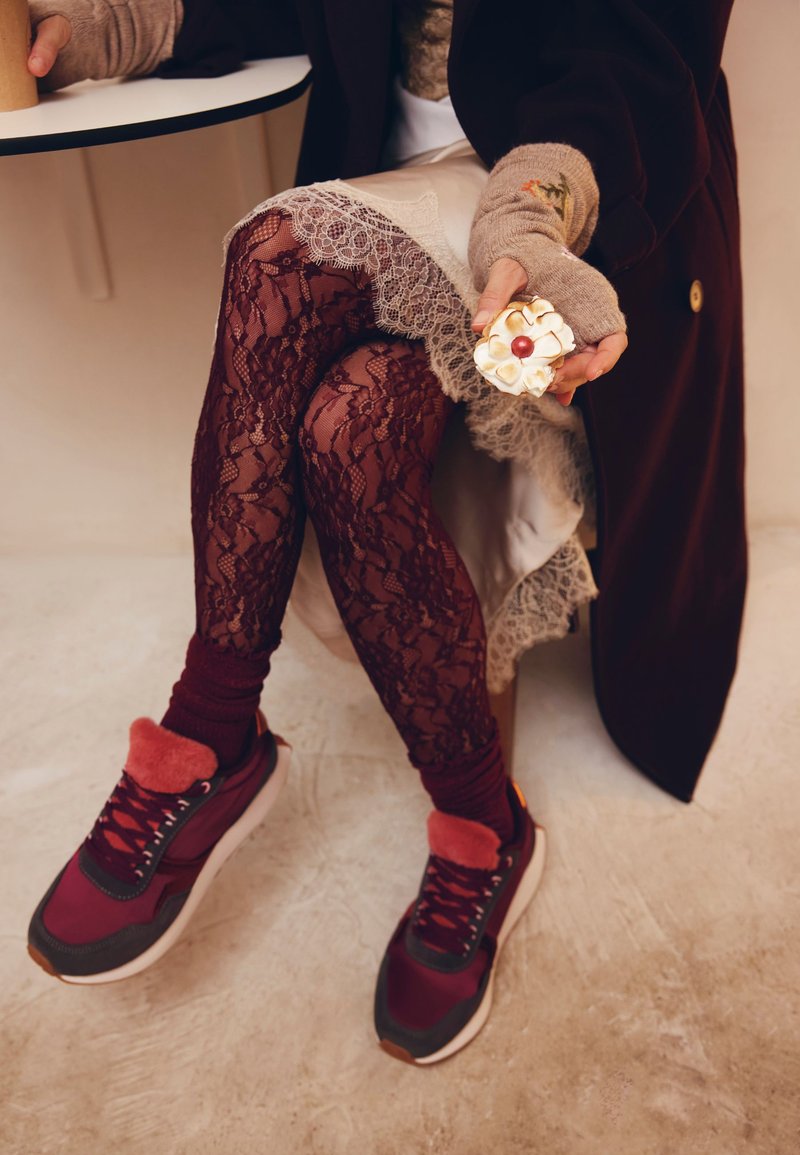 Chaussures de sport bordeaux avec des accents en fourrure rouge, associées à des leggings en dentelle noire. Une pâtisserie décorative est tenue dans une main. Sol clair.