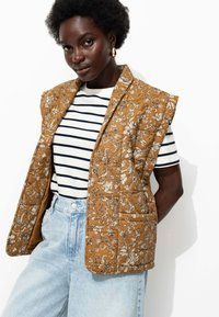 Gilet matelassé en moutarde avec un motif floral, associé à une chemise rayée. Le gilet a une coupe décontractée et des coutures contrastantes, ce qui accentue la texture.