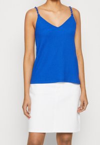 Modrý žebrovaný top na ramínka s výstřihem do V a ozdobnými ramínky, doplněný bílou sukní. Materiál vypadá hladký a lehký.