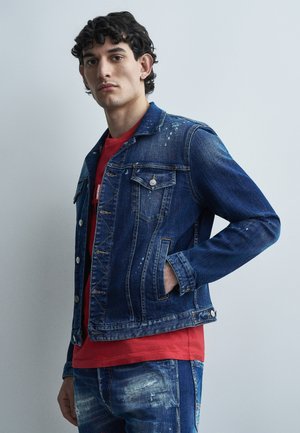Jeune homme aux cheveux bouclés et foncés portant une veste en jean bleue usée et un jean, avec un t-shirt rouge en dessous, la main dans la poche de la veste.