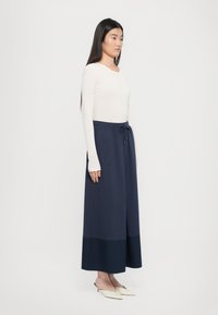 Helle beige Bluse mit langen Ärmeln, kombiniert mit einem dunkelblauen Midirock, der über einen Kordelzug in der Taille verfügt und einen kontrastierenden, dunkelblauen Saum hat.