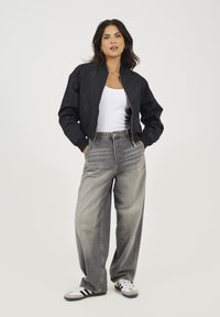 Czarna kurtka bomber, biała koszulka na ramiączkach, szare dżinsy o wysokim stanie oraz czarno-białe sneakersy. Modelka stoi z dłońmi w kieszeniach.