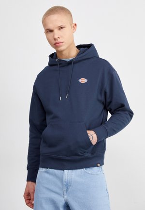 Felpa navy blu in materiale morbido con tasca frontale, cordini regolabili e patch con logo sul petto. Presenta polsini a coste.