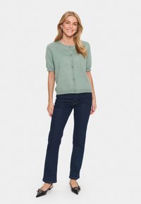 Ljusgrön kortärmad cardigan med rund halsringning och knappstängning, tillsammans med mörkblå raka jeans och svarta spetsiga skor.