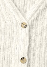 Nærbillede af hvid ribstrikket cardigan med to beige og brun marmoriserede knapper fastgjort langs foråbningen.