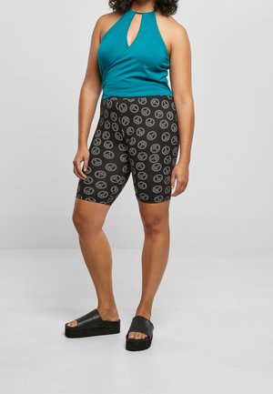 Femme portant un haut sans manches bleu sarcelle avec un col en trou de serrure, un short de cycliste noir avec des symboles de paix blancs et des sandales compensées noires.