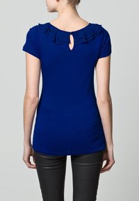 Blå kortärmad topp med volangdetalj vid halsringningen och nyckelhålsdetalj på baksidan. Tillverkad av ett mjukt tyg, figurnära design och rundad nederkant.